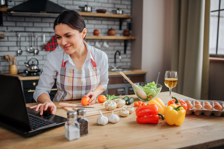 Cheerful adult woman sit at table in kitchen. She typing on keyboard and look on screen. Woman cook food